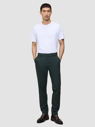 A man wearing a white t-shirt and dark green Ease Pants Regular Northern Pine with a tailored look, standing with his hands in his pockets.