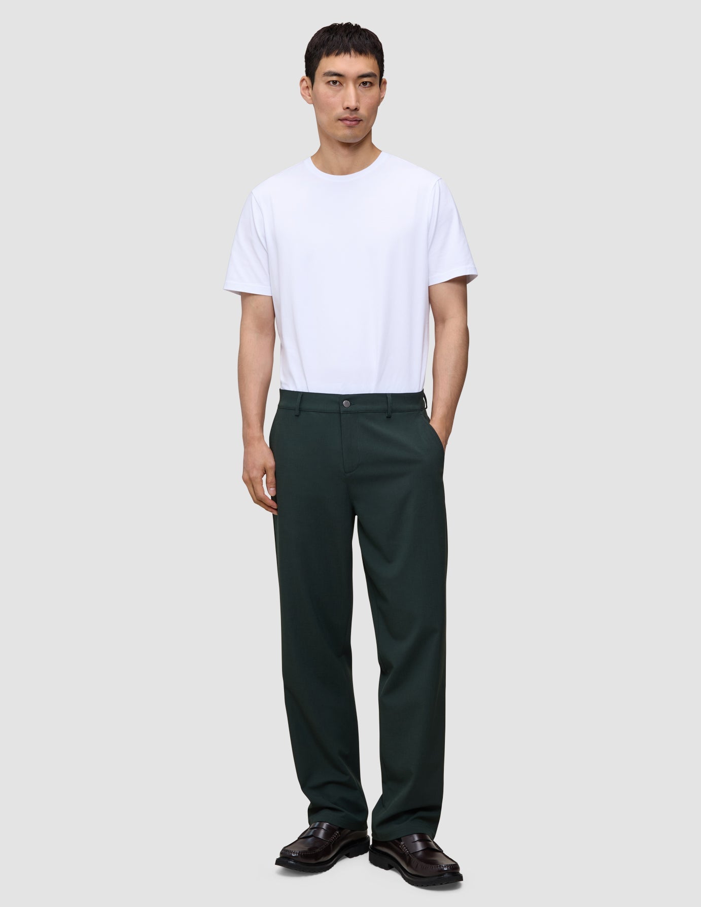A man wearing a white t-shirt, dark loafers, and Ease Pants Relaxed Fit Northern Pine, which are exceptionally soft, dark green relaxed-fit trousers.