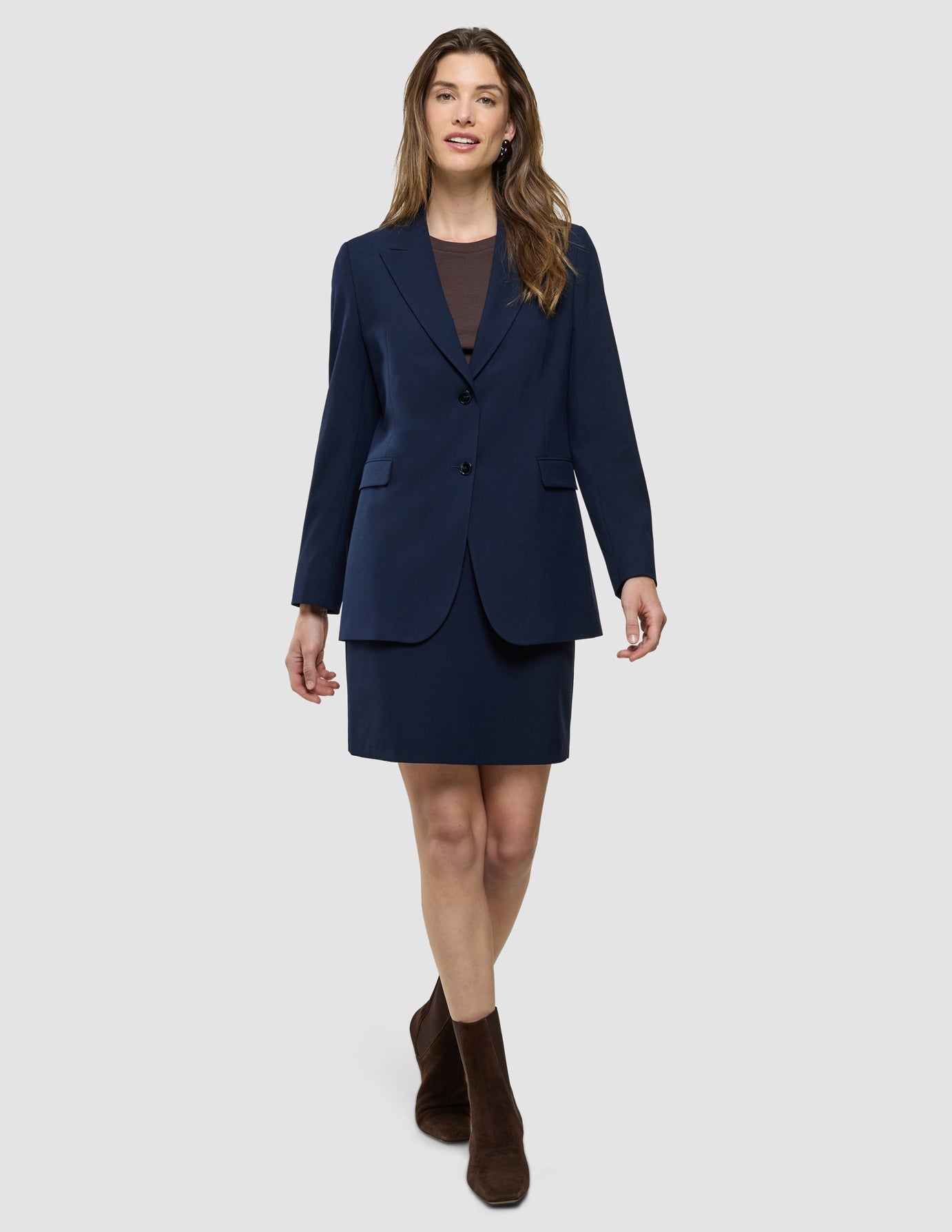 A smiling woman wearing the Ease Set Blue Nights Blazer and Short Skirt over a brown top with brown ankle boots.