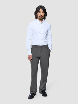 A man standing in a white button-down shirt, Ease Pants Relaxed Fit Dark Grey, and black loafers.