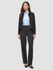 A woman wears an Essential Short Zip Jacket Black over a light blue collared shirt and dark grey trousers, paired with black block-heeled shoes.