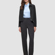 A woman wears an Essential Short Zip Jacket Black over a light blue collared shirt and dark grey trousers, paired with black block-heeled shoes.