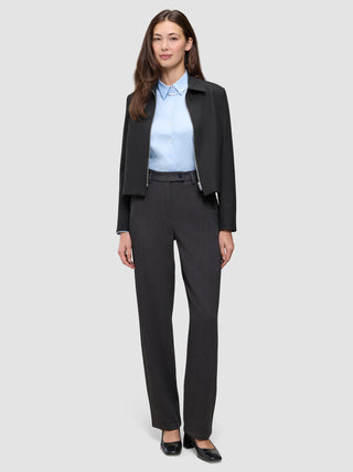 A woman wears an Essential Short Zip Jacket Black over a light blue collared shirt and dark grey trousers, paired with black block-heeled shoes.