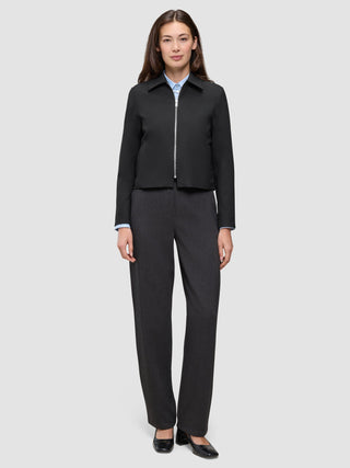 A woman wearing an Essential Short Zip Jacket Black over a light blue collared shirt, paired with charcoal grey trousers and black heels.