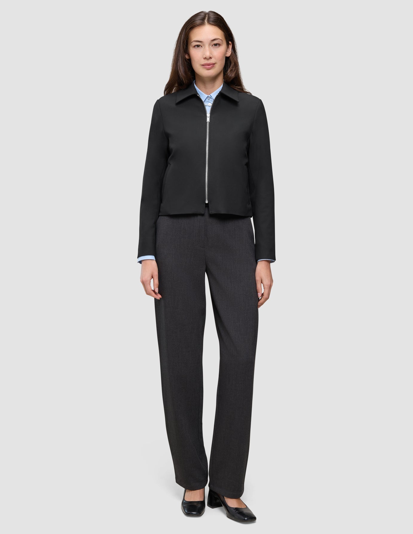 A woman wearing an Essential Short Zip Jacket Black over a light blue collared shirt, paired with charcoal grey trousers and black heels.