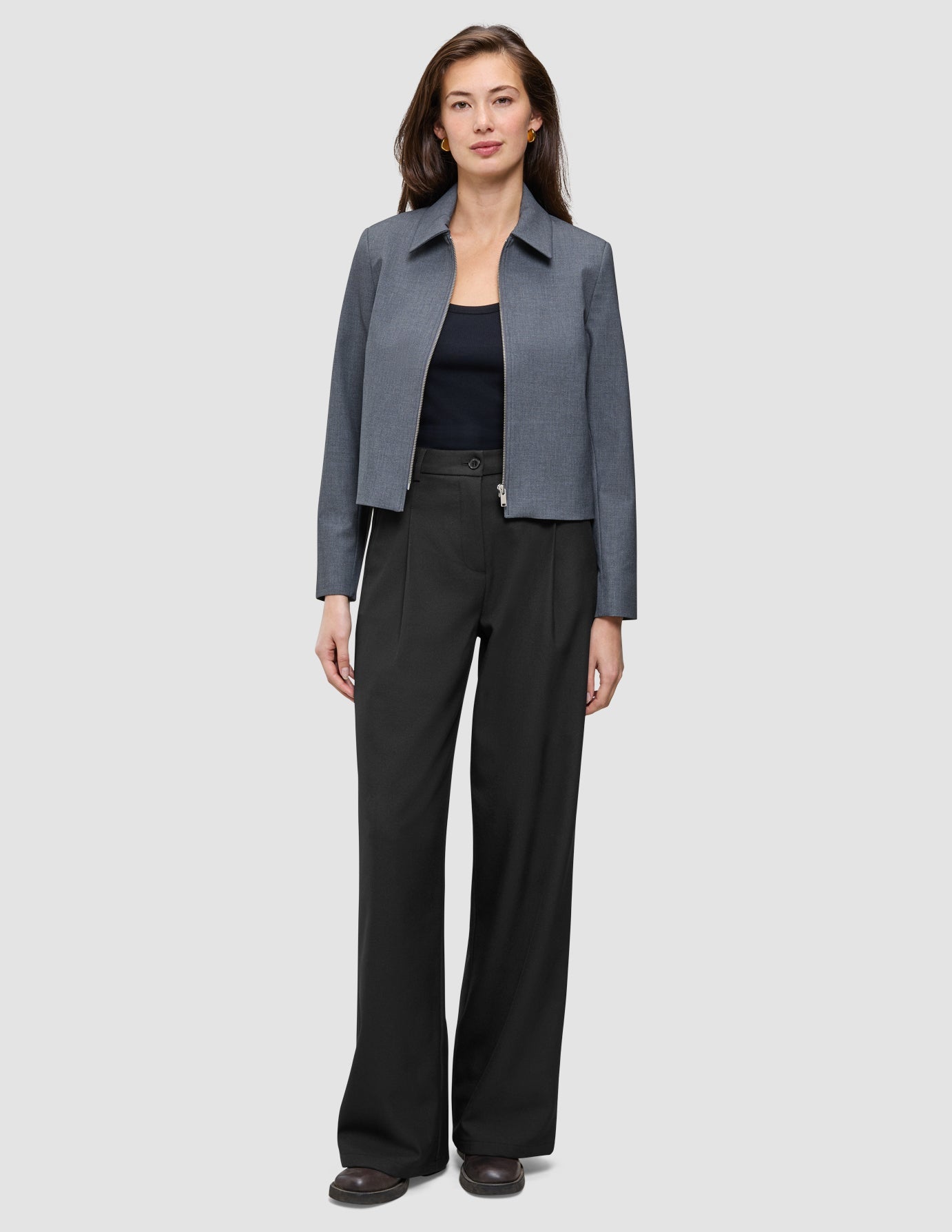 A woman wears a dark grey melange Essential Short Zip Jacket over a black top and wide-leg trousers.