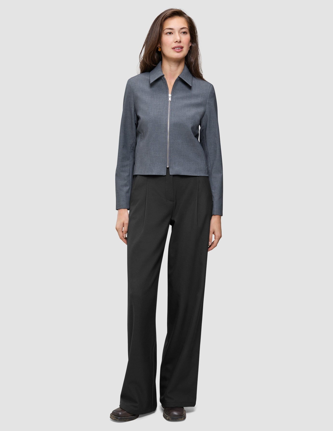 A model wearing an Essential Short Zip Jacket Dark Grey Melange with a minimalist collared zip-front and wide-leg black trousers.
