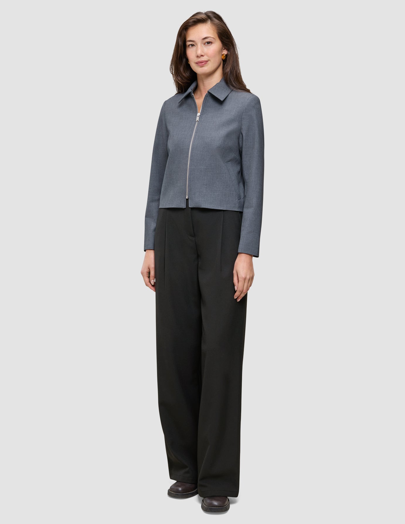 A woman wearing an Essential Short Zip Jacket Dark Grey Melange and loose black trousers.