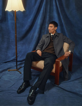 A man wearing a Maxi Wool Coat Dark Grey over a brown sweater and dark trousers sits in a wooden armchair next to a floor lamp.