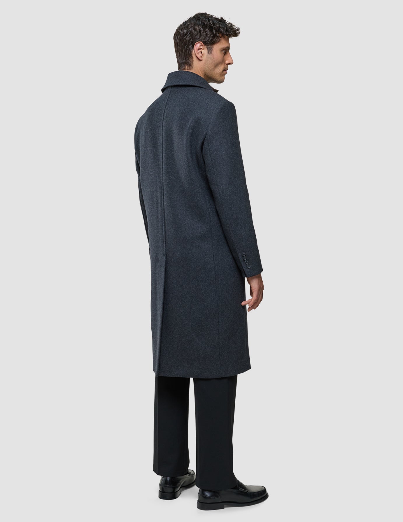 A man viewed from behind, wearing a knee-length Maxi Wool Coat Dark Grey over black trousers and black dress shoes.