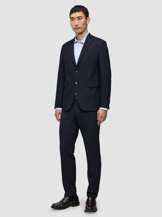 A man wears a Motion Blazer Regular Midnight Blue with matching trousers, a light blue button-down shirt, and brown loafers, all crafted from breathable fabric.
