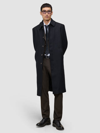 A man wears a black overcoat over a suit, white shirt, and tie, paired with Motion Suit Pants Regular Cognac and black dress shoes.