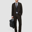 A man wearing Motion Suit Pants Regular Cognac and a matching breathable jacket with a white dress shirt and dark tie, holding a black backpack.