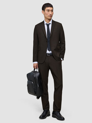 A man wearing Motion Suit Pants Regular Cognac and a matching breathable jacket with a white dress shirt and dark tie, holding a black backpack.