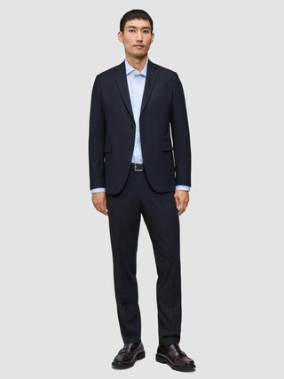 A man wearing Motion Suit Pants Regular Midnight Blue with a matching lightweight navy suit jacket, a light blue button-down shirt, and dark leather loafers.