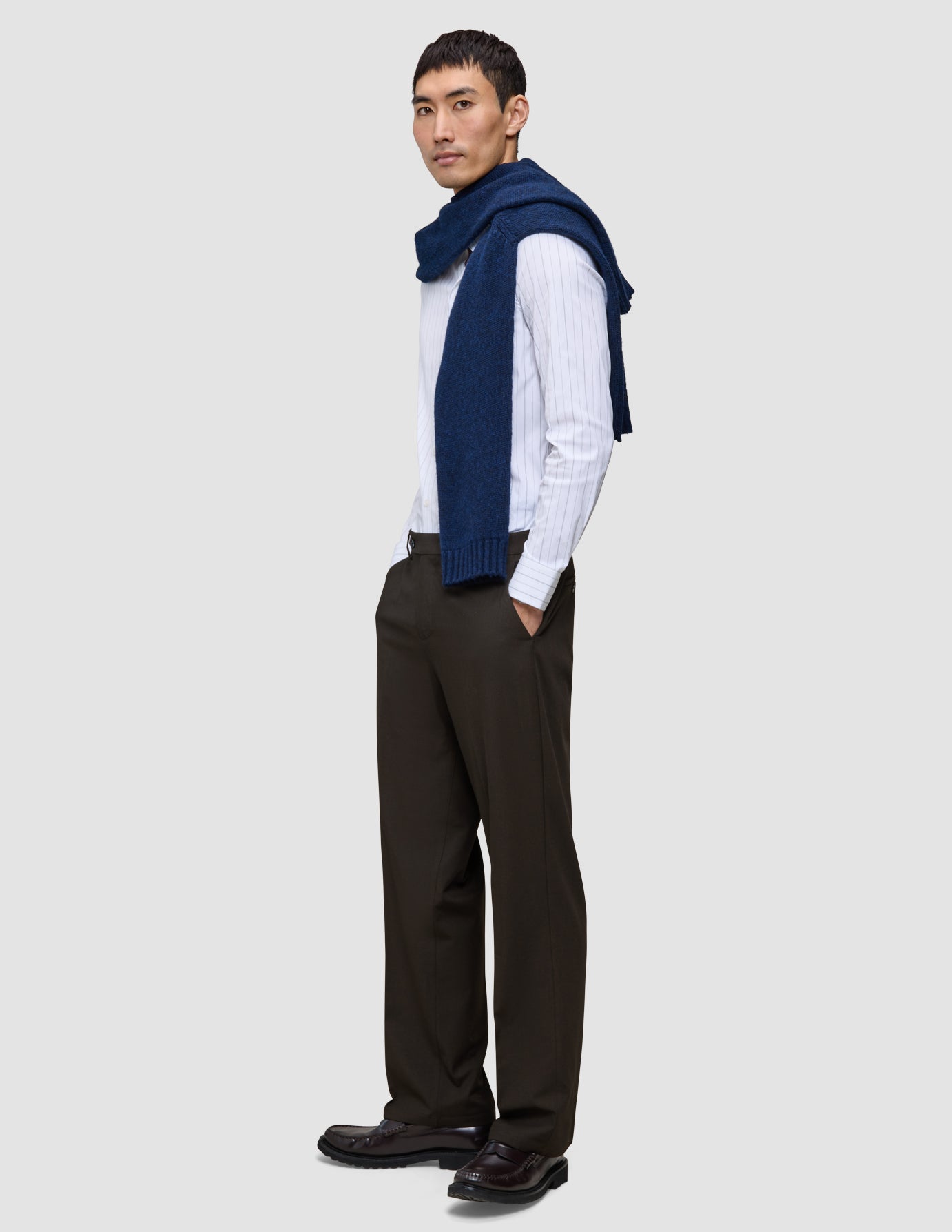 A man wearing a white pinstriped button-down shirt, a navy blue sweater draped over his shoulders, loafers, and Motion Suit Pants Relaxed Cognac.