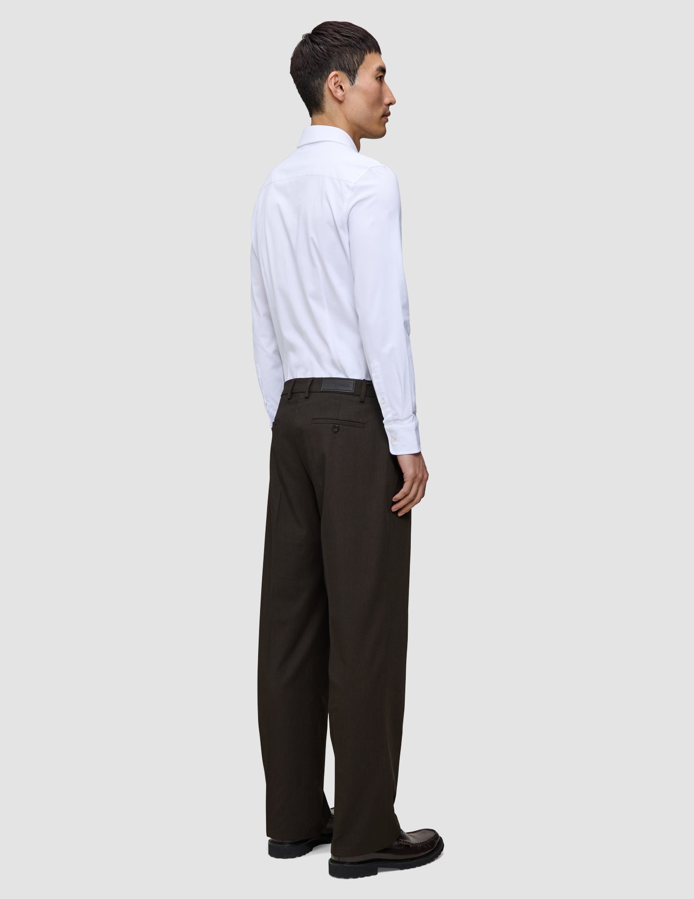 A man viewed from behind wears a white button-down shirt, Motion Suit Pants Relaxed Cognac, and dark brown loafers.