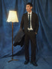 Man wearing a brown Motion Suit Pants Relaxed Cognac, a white dress shirt, and a dark tie, holding a jacket while standing next to a floor lamp.