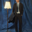 Man wearing a brown Motion Suit Pants Relaxed Cognac, a white dress shirt, and a dark tie, holding a jacket while standing next to a floor lamp.