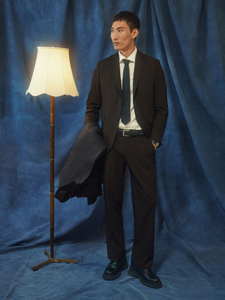 Man wearing a brown Motion Suit Pants Relaxed Cognac, a white dress shirt, and a dark tie, holding a jacket while standing next to a floor lamp.