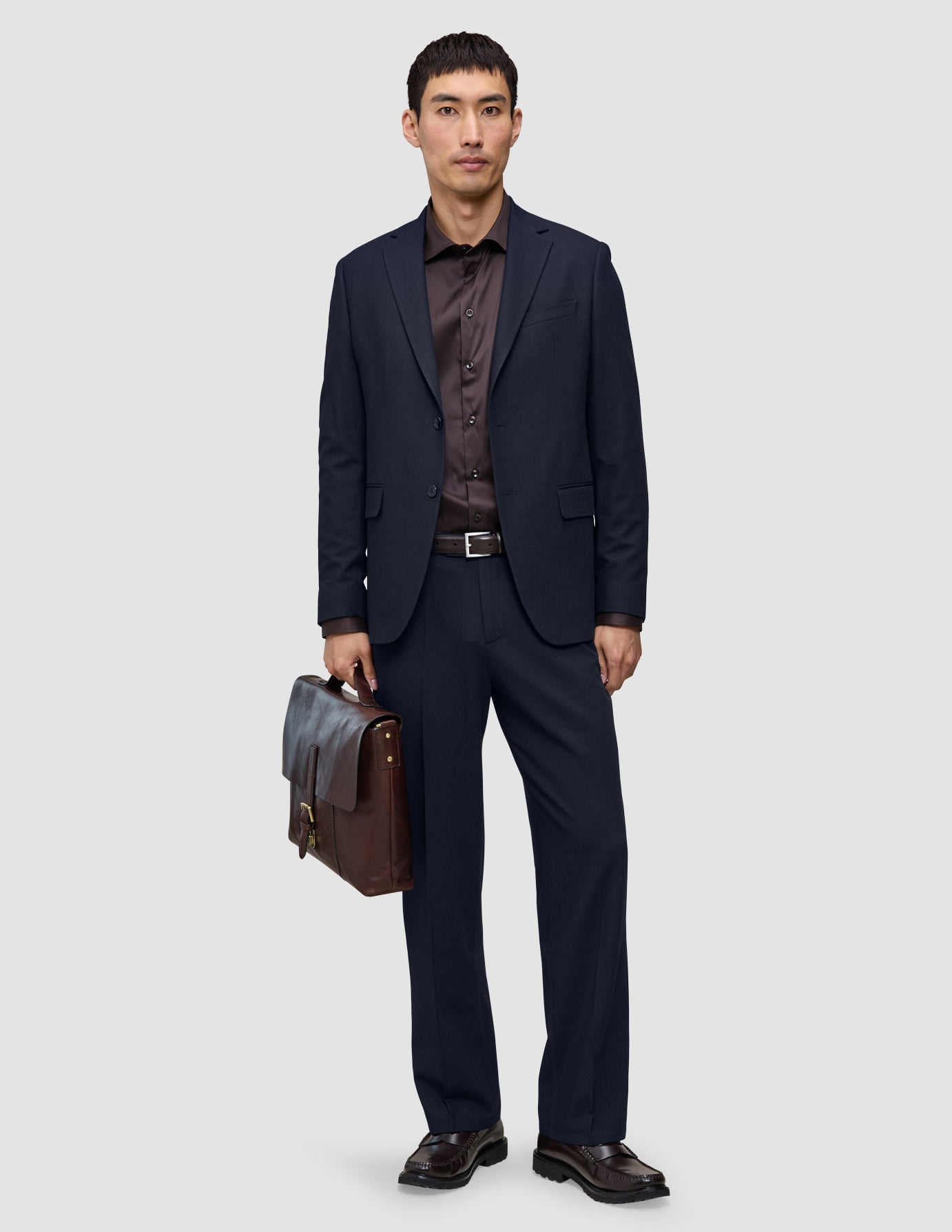 A man holding a brown leather briefcase, wearing a dark blue suit jacket, a dark brown button-down shirt, dark loafers, and Motion Suit Pants Relaxed Midnight Blue.