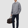A man wearing a grey crewneck sweater over a white collared shirt and midnight blue Motion Suit Pants Slim, carrying a brown duffel bag and wearing dark loafers.