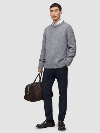 A man wearing a grey crewneck sweater over a white collared shirt and midnight blue Motion Suit Pants Slim, carrying a brown duffel bag and wearing dark loafers.