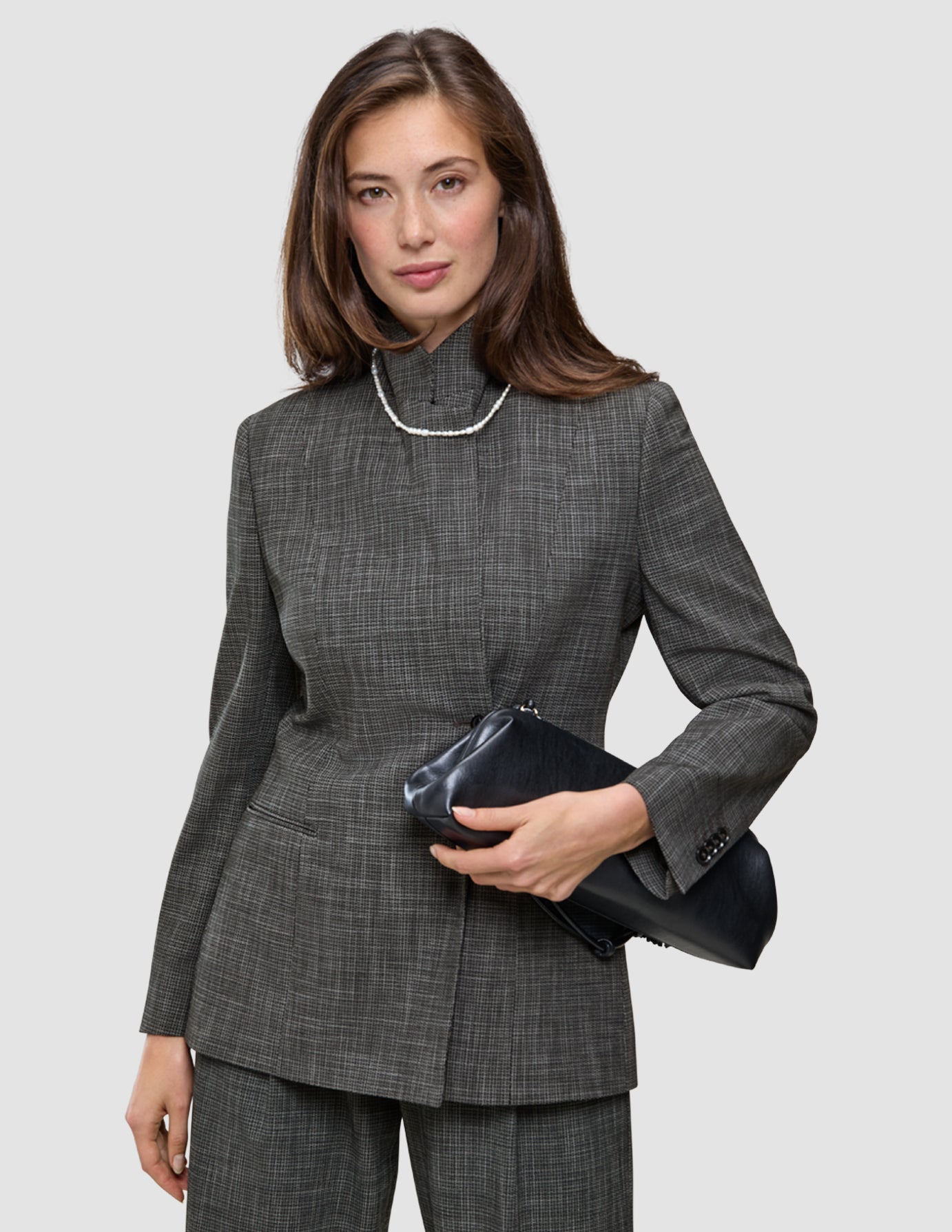 A woman wears The suit with a statement in grey plaid, featuring a blazer and matching wide-leg pants paired with a high-neck jacket, a single-strand pearl necklace, and a black clutch.
