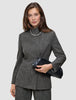 A woman wears The suit with a statement in grey plaid, featuring a blazer and matching wide-leg pants paired with a high-neck jacket, a single-strand pearl necklace, and a black clutch.