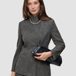 A woman wears The suit with a statement in grey plaid, featuring a blazer and matching wide-leg pants paired with a high-neck jacket, a single-strand pearl necklace, and a black clutch.