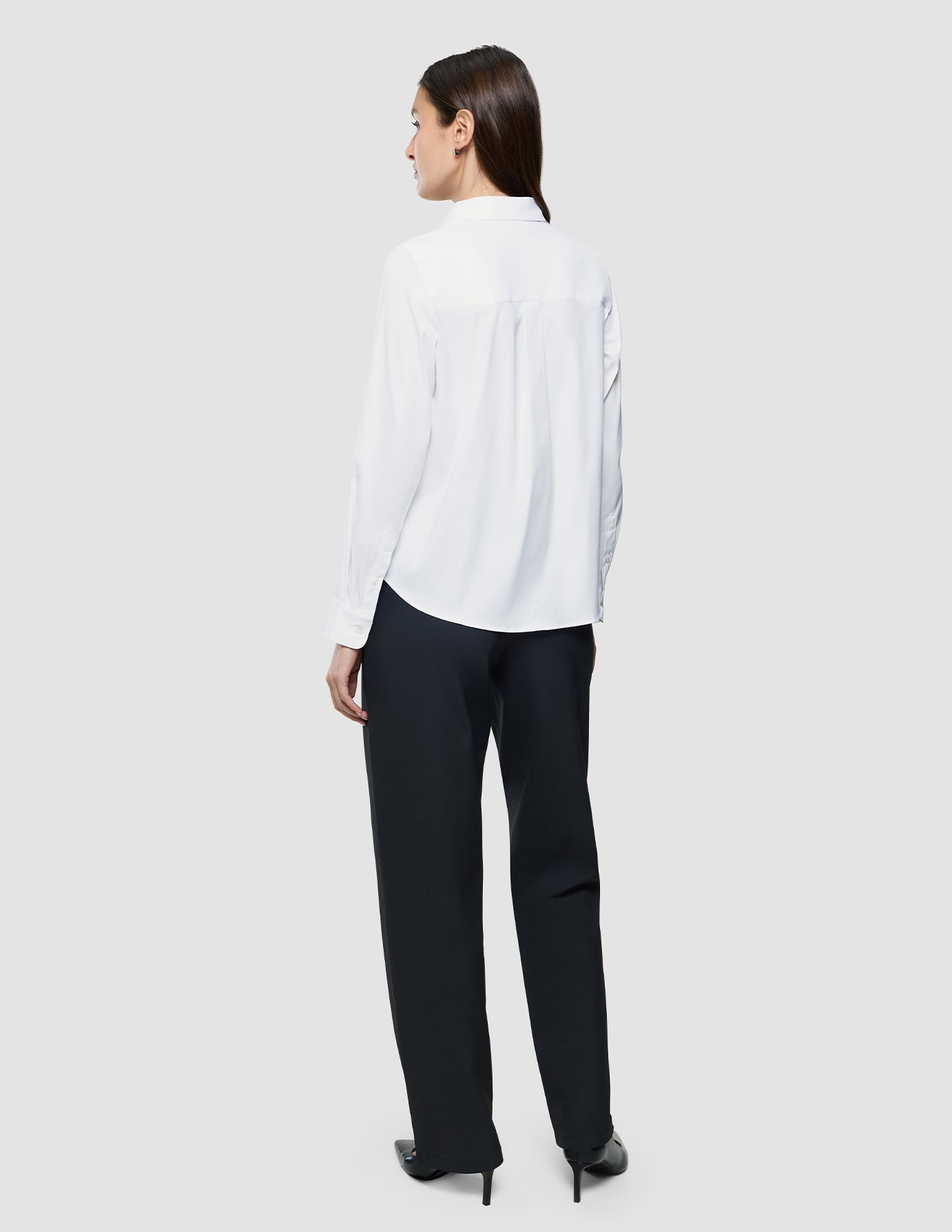 A woman stands facing away in a Women Oxford Shirt White Regular and black trousers with heels, set against a light gray background, evoking minimalist Scandinavian style.