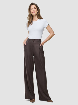 A woman wearing a white t-shirt and Women Raina Pants Wide Chestnut Pinstripe with her hands in her pockets, styled with a Scandinavian design aesthetic.