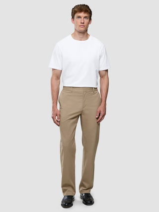 A man wearing a white t-shirt, Refine Cotton Pants Relaxed Fit Sand, and black dress shoes stands against a light gray background.