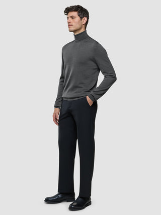 A man wearing a Seamless Premium Merino Knit Turtleneck Charcoal Grey and black trousers, standing with his hand in his pocket.