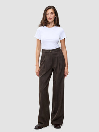 A person wearing a white t-shirt and Serene Pants Wide Walnut Herringbone, which are high-waisted, dark brown pinstriped trousers with a wide-leg silhouette.