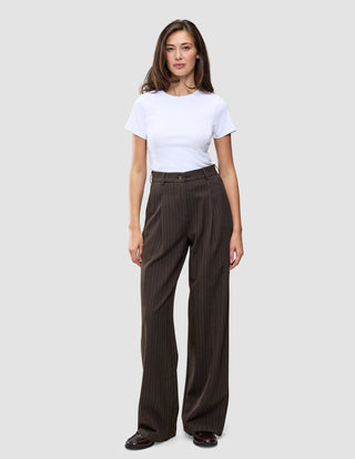 A person wearing a white t-shirt and Serene Pants Wide Walnut Herringbone, which are high-waisted, dark brown pinstriped trousers with a wide-leg silhouette.