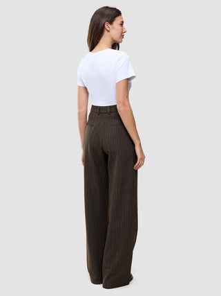 A woman wearing a white t-shirt and Serene Pants Wide Walnut Herringbone, high-waisted, dark brown, pinstriped, wide-leg trousers, viewed from the back.