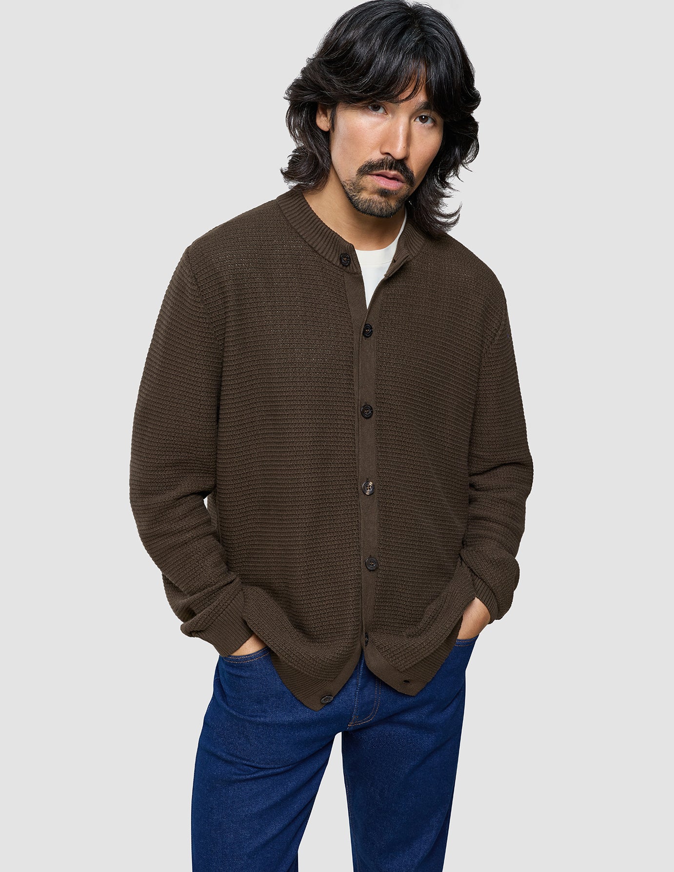 Man wearing a brown textured cardigan over a white shirt with blue pants on a light gray background