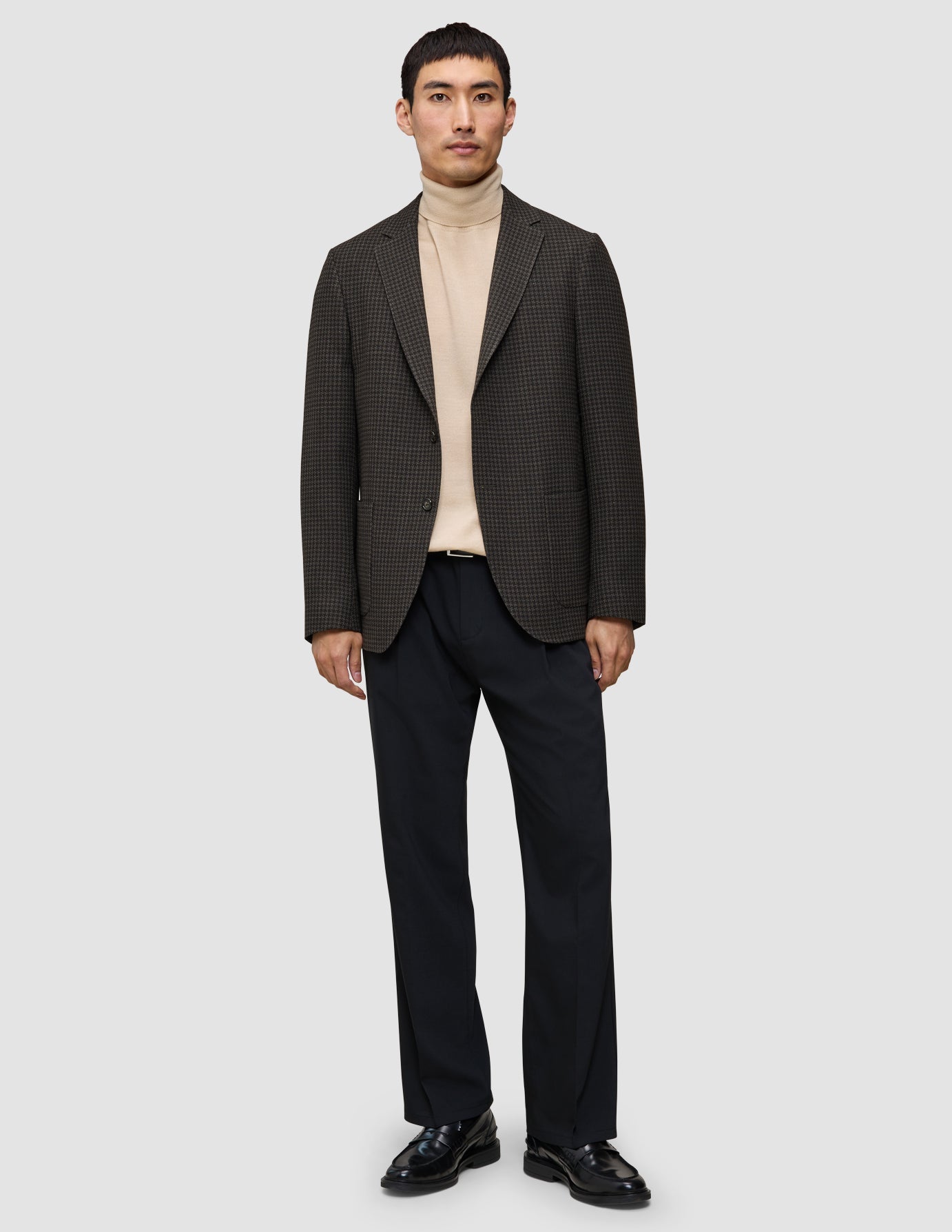 A man wearing The knit-and-blazer look, consisting of a tan merino crewneck, a dark patterned wool blazer, black trousers, and black loafers.
