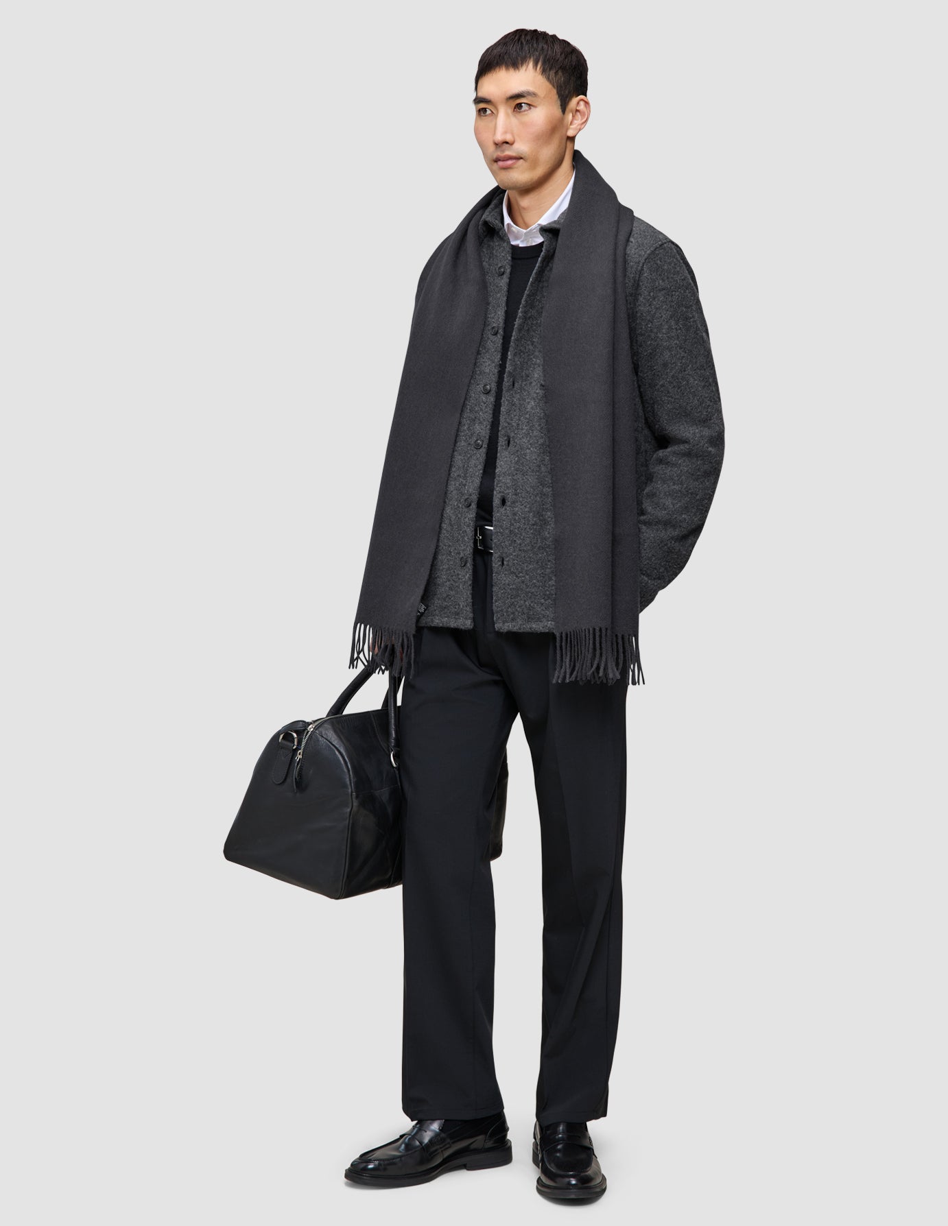 A man wearing Layers that work, featuring a double layer wool overshirt over a premium merino knit and white shirt, a dark scarf, black trousers, and loafers, while holding a black duffel bag.