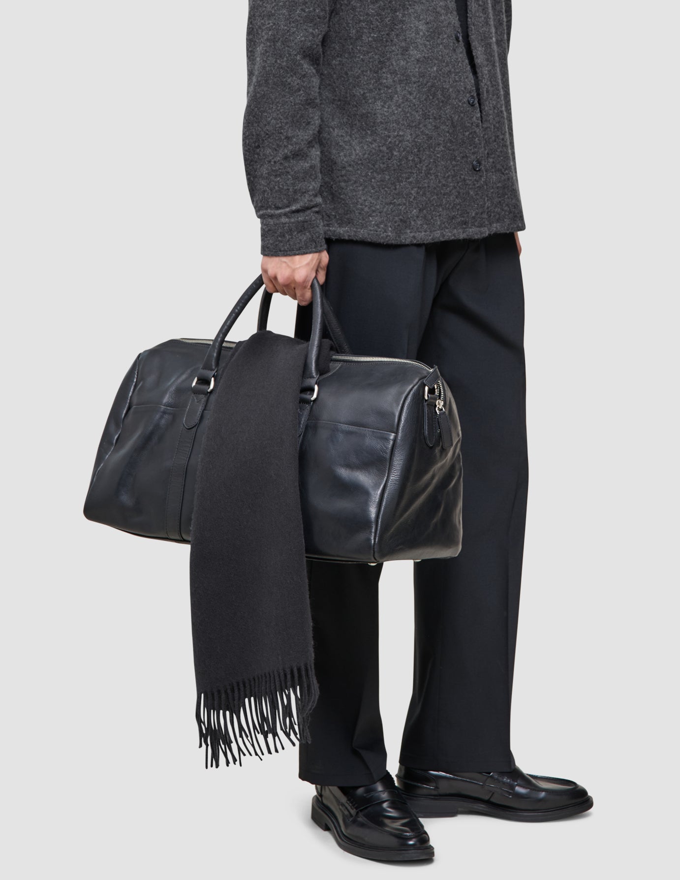 A person wearing Layers that work, featuring a premium merino knit sweater and tech wool pants, holds a black leather duffel bag with a black scarf draped over the handle.