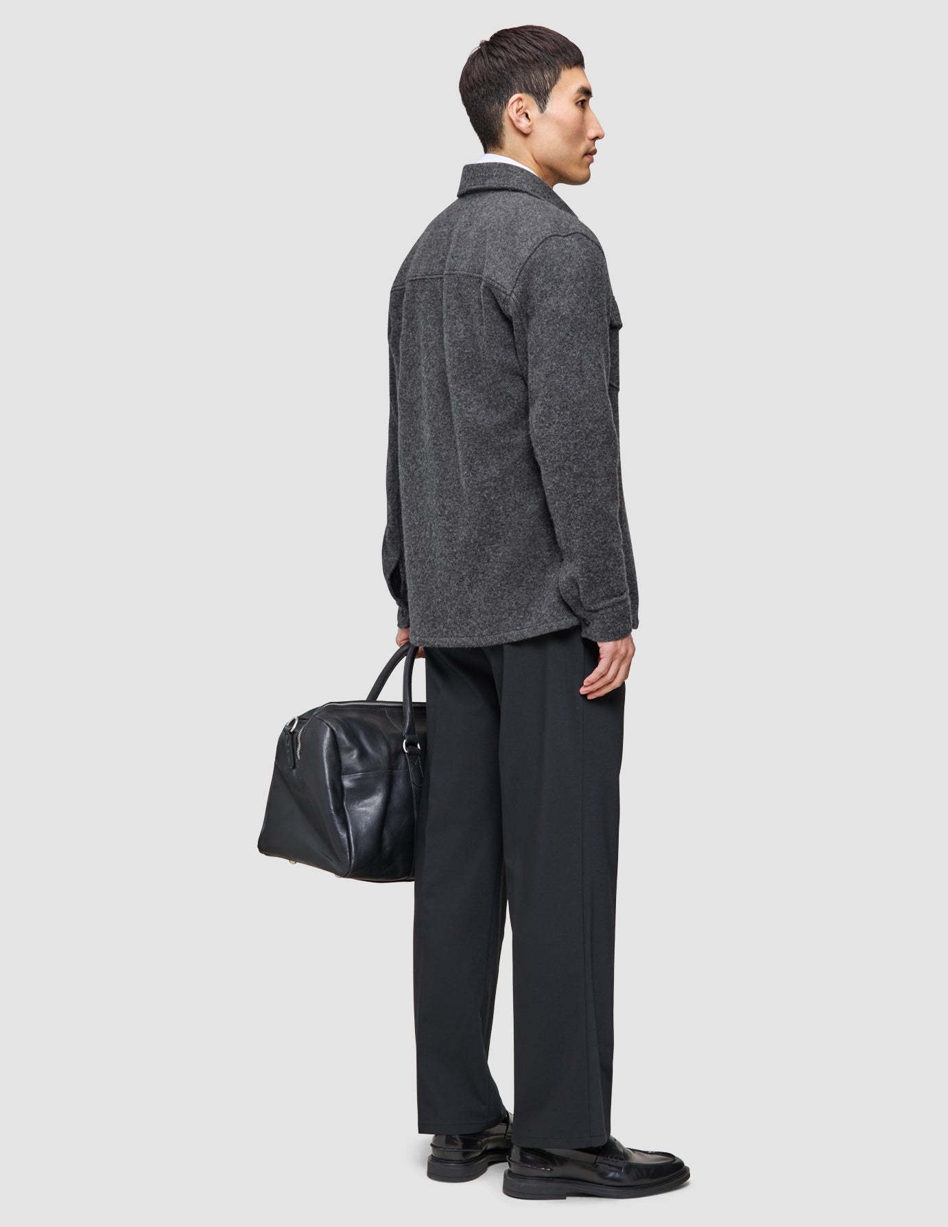 A man wearing Layers that work, featuring a double layer wool overshirt and tech wool pants, carries a black leather duffel bag.