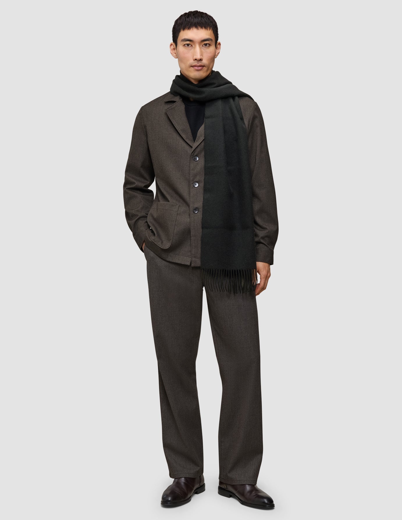 A man wearing A different kind of suit consisting of a dark brown Atelier Overshirt and matching Atelier Pants, a black turtleneck, and a long black scarf, paired with dark brown leather shoes.