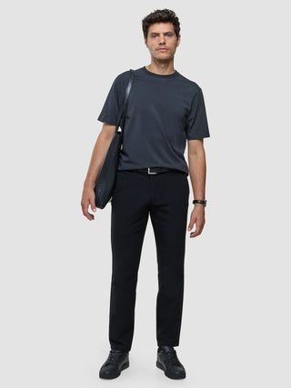 A man in a Supima T-shirt Box Fit Deep Grey and black trousers with a shoulder bag stands against a plain background, showcasing a clean Scandinavian design.