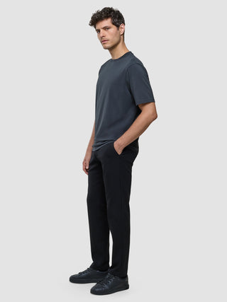 A man wearing a Supima T-shirt Box Fit Deep Grey, black trousers, and black sneakers stands with his hand in his pocket, showcasing a clean Scandinavian design.