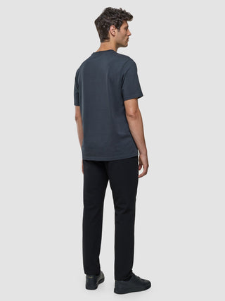 A man standing and facing away, wearing a Supima T-shirt Box Fit Deep Grey, black trousers, and black sneakers in a clean, Scandinavian design.