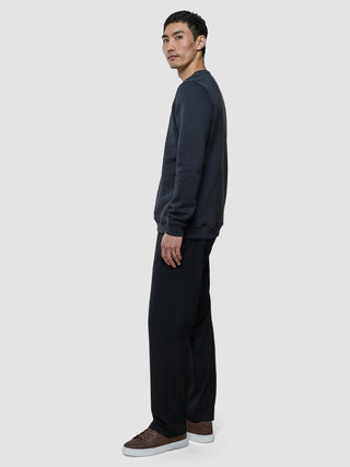A man stands in profile wearing a Sweat Crewneck Deep Grey, black trousers, and brown sneakers.
