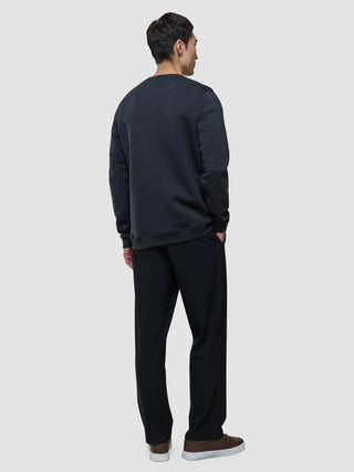 A person seen from behind wearing a Sweat Crewneck Deep Grey, black trousers, and brown sneakers.