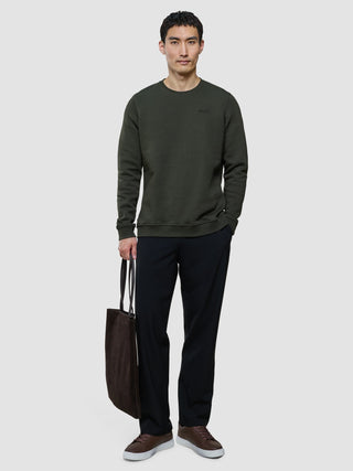 A man stands wearing a Sweat Crewneck Green Night and dark trousers, holding a dark brown tote bag.