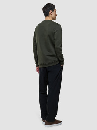 A man standing with his back to the camera, wearing a Sweat Crewneck Green Night, black trousers, and brown sneakers.